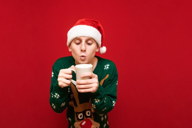 homme en bonnet de noel et pull vert se tient avec une tasse de boisson chaude dans les mains sur fond rouge