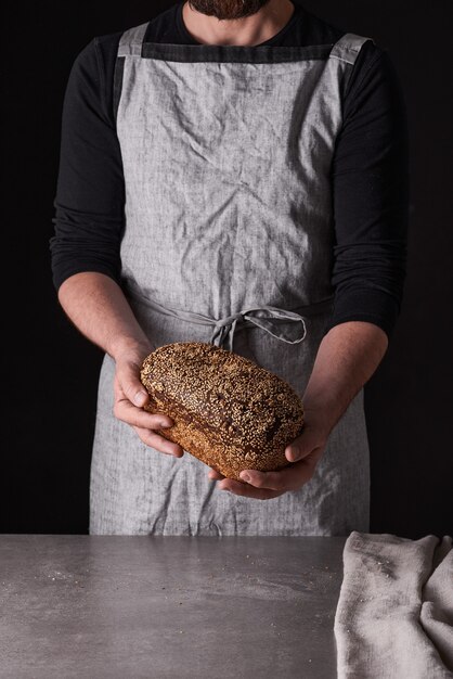 Un homme boulanger avec une barbe dans un tablier gris se dresse sur un fond noir et tient, casse, coupe un délicieux pain croustillant, des petits pains, de la baguette.