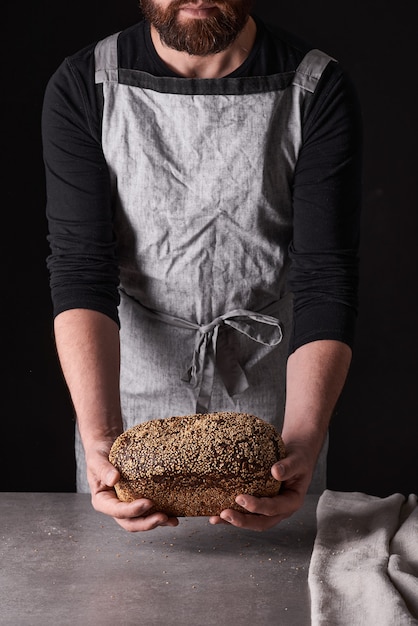 Un homme boulanger avec une barbe dans un tablier gris se dresse sur un fond noir et tient, casse, coupe un délicieux pain croustillant, des petits pains, de la baguette.