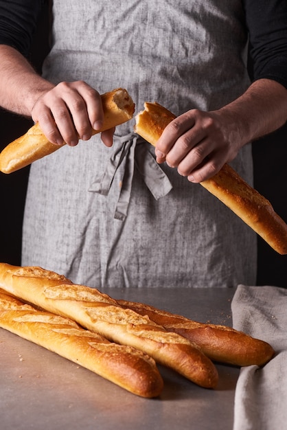 Un homme boulanger avec une barbe dans un tablier gris se dresse sur un fond noir et tient, casse, coupe un délicieux pain croustillant, des petits pains, de la baguette.