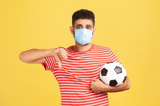 Homme bouleversé dans un masque de sécurité médicale et en t-shirt rouge tenant une balle montrant qu'il n'aime pas l'annulation du championnat en raison d'une épidémie de coronavirus Tourné en studio intérieur isolé sur fond jaune