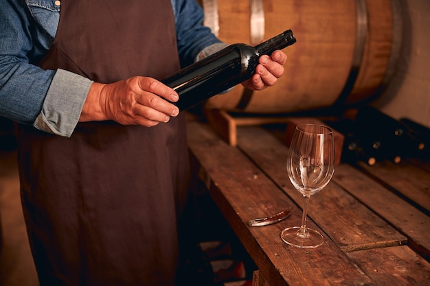 homme avec une bouteille de boisson alcoolisée dans ses mains debout près d'une table en bois avec un verre à vin dans une cave à vin