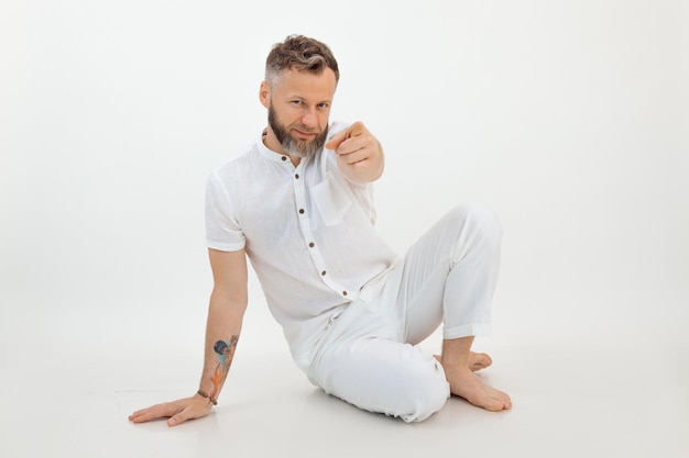 Homme branché barbu avec tatouage assis sur le sol sur fond blanc pointant la caméra par l'index