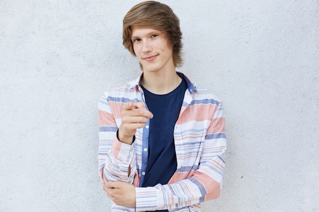 Homme branché avec coupe de cheveux élégante portant une chemise décontractée pointant avec le doigt à la caméra ayant un look timide isolé sur fond blanc. Adolescent, choisir quelque chose en faisant des gestes avec le doigt