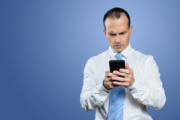 Homme brésilien d'âge mûr, dans des vêtements formels, tenant son smartphone avec un visage sérieux. Fond bleu.