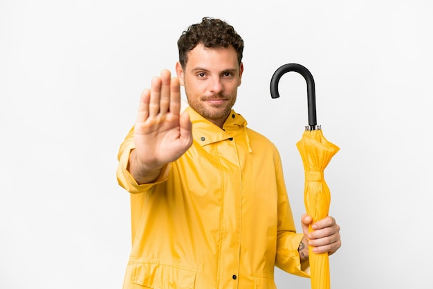 Homme brésilien avec manteau imperméable et parapluie sur fond blanc isolé faisant un geste d'arrêt