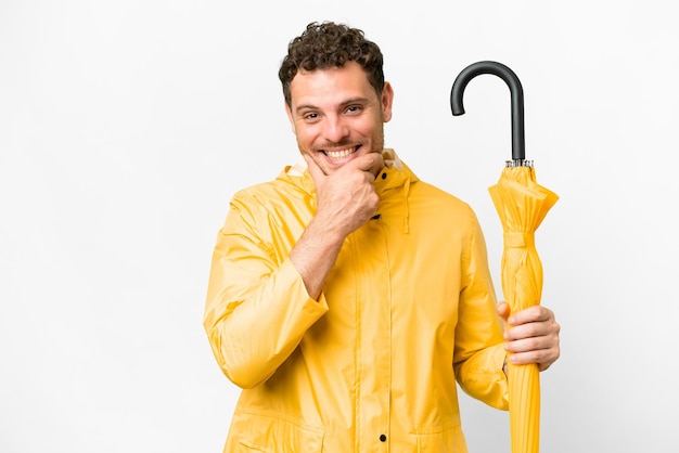 Homme brésilien avec manteau imperméable et parapluie sur fond blanc isolé heureux et souriant