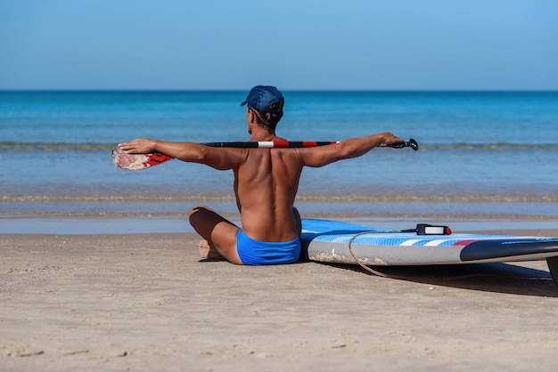 Homme bronzé est assis sur la plage avec une planche de surf et une rame et regarde vers la mer.