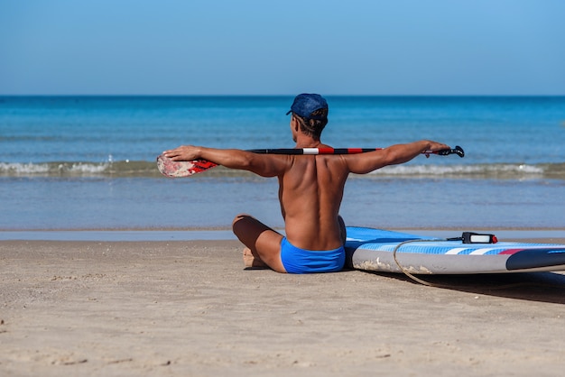 Homme bronzé est assis sur la plage avec une planche de surf et une rame et regarde vers la mer.