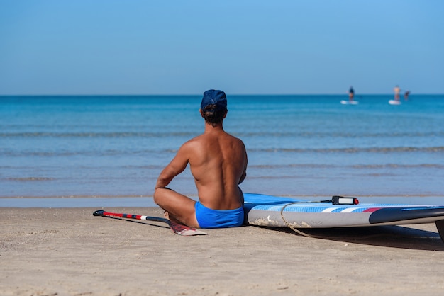 Un homme bronzé est assis sur la plage près de sa planche de surf et regarde la mer.