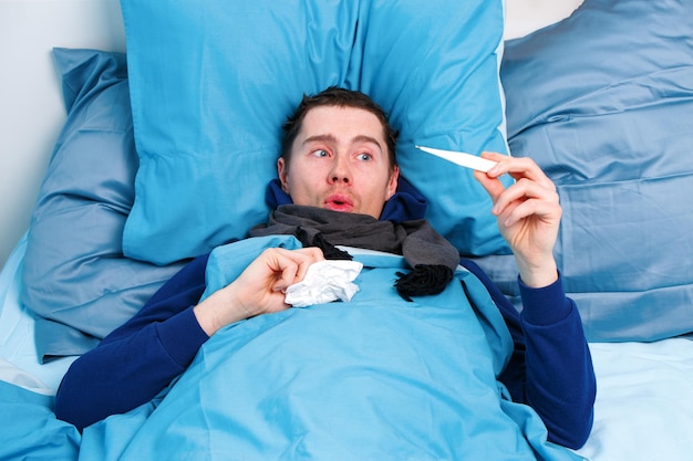 Homme brun malade en écharpe avec thermomètre à la main couché dans son lit
