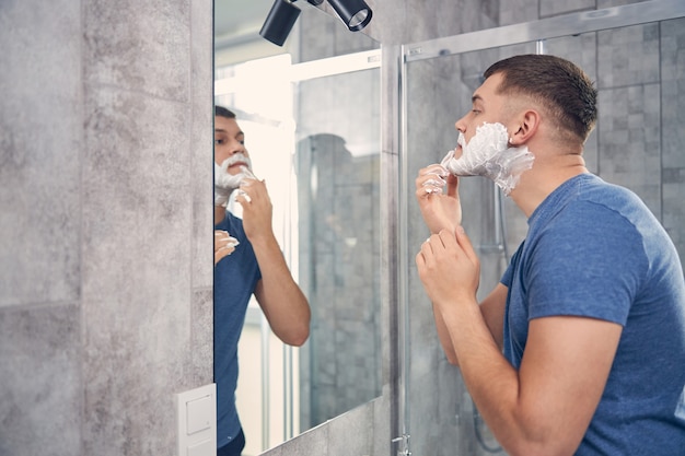 Homme brune séduisante en T-shirt bleu passant du temps dans la salle de bain devant le miroir tout en se rasant le visage