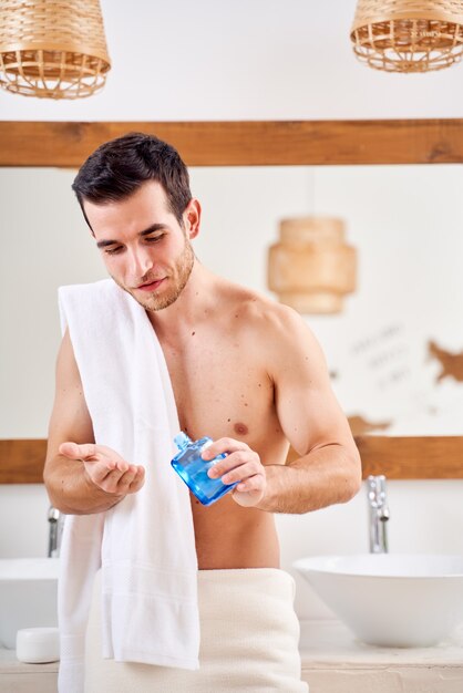 L'homme Brunet avec une serviette sur ses épaules verse de la lotion dans sa main devant le miroir de la salle de bain