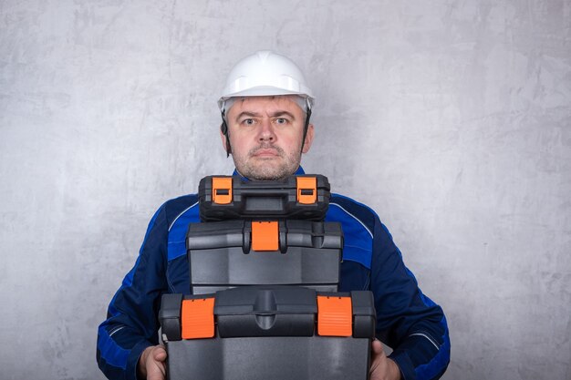 Un homme brutal dans un uniforme de travail et un casque blanc tient des boîtes d'outils dans ses mains. l'image isolée sur fond gris