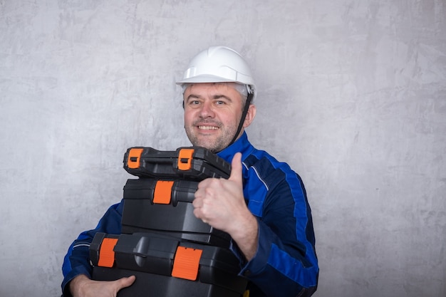 Un homme brutal dans un uniforme de travail et un casque blanc tient des boîtes avec des outils dans ses mains et montre un geste comme. l'image isolée sur fond gris