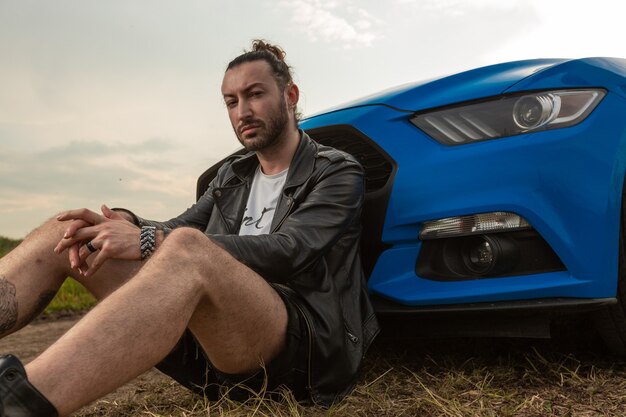 Homme brutal en veste de cuir est assis appuyé sur une voiture de sport bleu vif au milieu du terrain photo de haute qualité