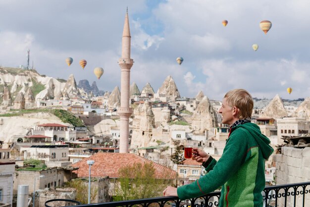 Homme buvant un verre de thé turc en face de la vieille ville de Göreme en Cappadoce en regardant la montgolfière. Belle journée ensoleillée et vue sur la mosquée depuis la terrasse.