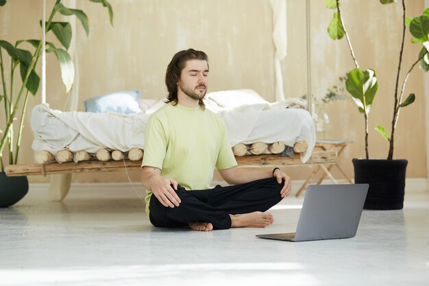 Homme calme et détendu en posture de lotus avec ordinateur portable sur le sol, beau mâle en chemise verte assis en méditation et en se concentrant