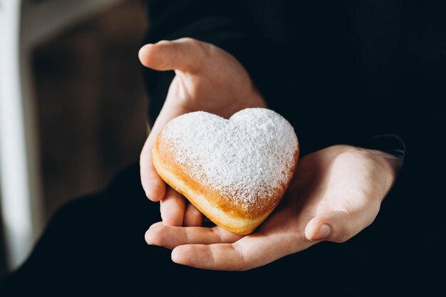 Un homme avec une capuche noire tient un donut en forme de cœur recouvert de sucre en poudre