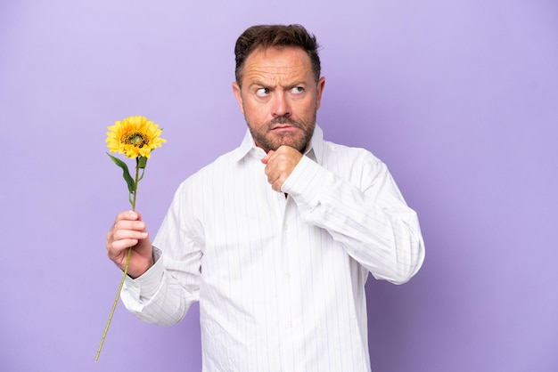 Homme caucasien d'âge moyen tenant une fleur de soleil isolée sur fond violet ayant des doutes et pensant