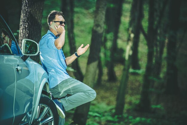 Photo un homme caucasien ayant une conversation téléphonique tendue gesticulant nerveusement et appuyant son pied contre la roue de sa voiture argentée forêt profonde à l'arrière-plan