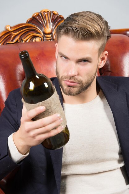 Homme caucasien avec barbe, cheveux élégants tenir une bouteille de vin sur un fauteuil en cuir marron.