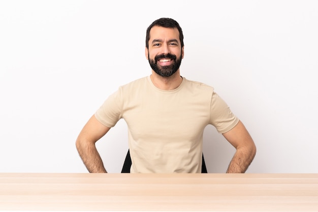 Homme caucasien avec barbe dans une table posant avec les bras à la hanche et souriant.