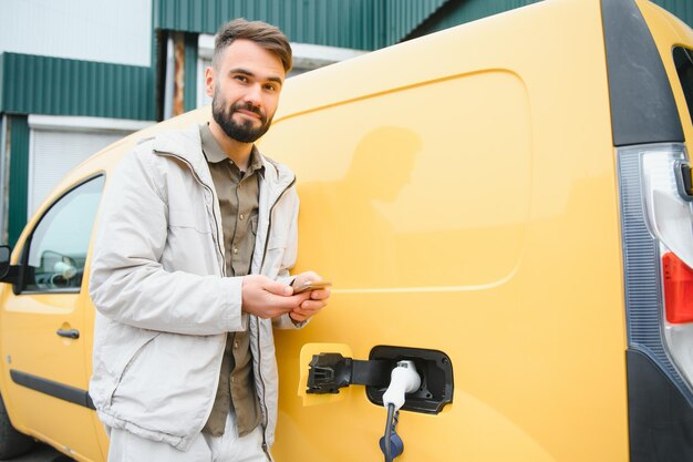 Homme caucasien barbu debout près d'une voiture électrique qui charge et fait des ajustements de temps sur un smartphone