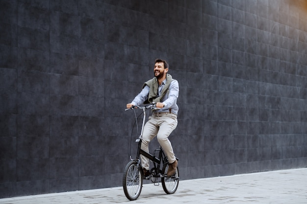 Homme caucasien élégant barbu caucasien souriant, écouter de la musique et faire du vélo. En arrière-plan, un mur gris.