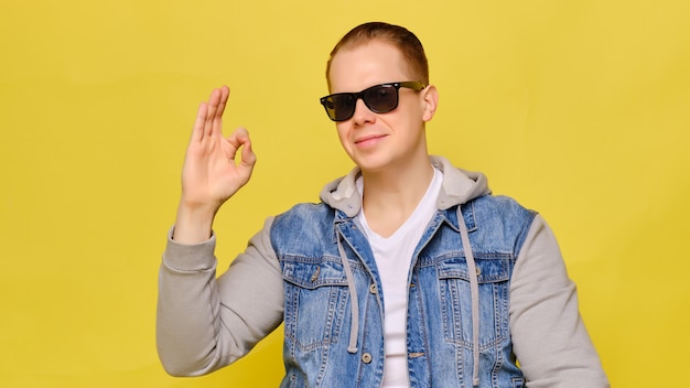 Homme caucasien élégant dans un jean et des lunettes de soleil sur fond jaune. La main montre abstraitement que tout va bien.