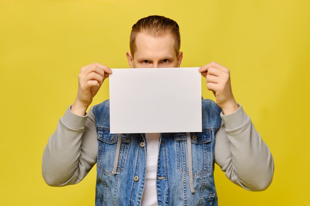 Homme caucasien élégant en jeans sur fond jaune. Mains tenant une feuille de papier blanc et couvrant la moitié du visage.