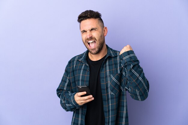 Homme caucasien isolé sur fond violet avec téléphone en position de victoire