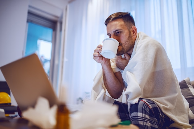 Homme caucasien mal rasé malade en pyjama recouvert de couverture assis sur un canapé dans le salon, boire du thé et regarder un ordinateur portable.