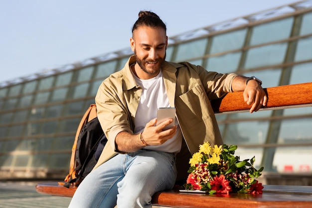 Un homme caucasien millénaire positif avec un bouquet attendant sa femme passe un appel vidéo sur son smartphone