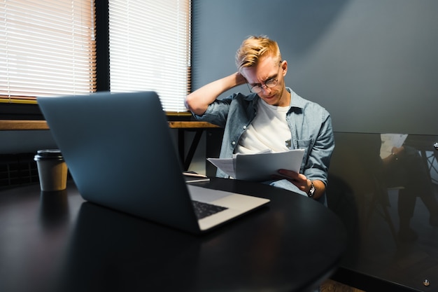 Homme caucasien pensif travaillant avec un ordinateur portable au bureau à domicile et à la recherche de documents papier, calcul des factures