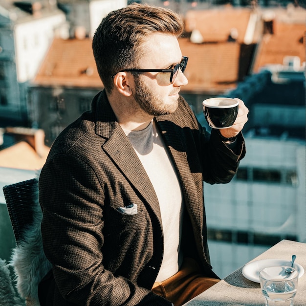 Un homme caucasien portant des lunettes boit du café dans une tasse au café. Travail à distance. La vie en ville. Homme d'affaires à la pause. Mode casual. Notion de réussite. Adulte souriant attrayant. Loisirs. Technique mixte.