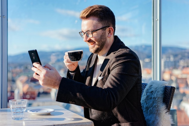Un homme caucasien portant des lunettes tient un smartphone et boit du café dans une tasse au café. Travail à distance via smartphone. La vie en ville. Homme d'affaires à la pause. Mode casual. Notion de réussite. Technique mixte.