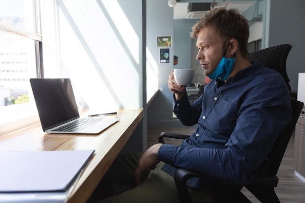 Photo homme caucasien portant un masque dans un bureau. assis, utilisant un ordinateur portable et buvant une tasse de café. hygiène sur le lieu de travail pendant la pandémie du coronavirus covid 19.