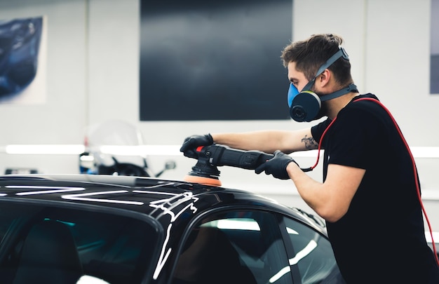 Homme caucasien portant un masque de protection polissant la peinture de voiture noire à l'aide d'une voiture-outil professionnelle