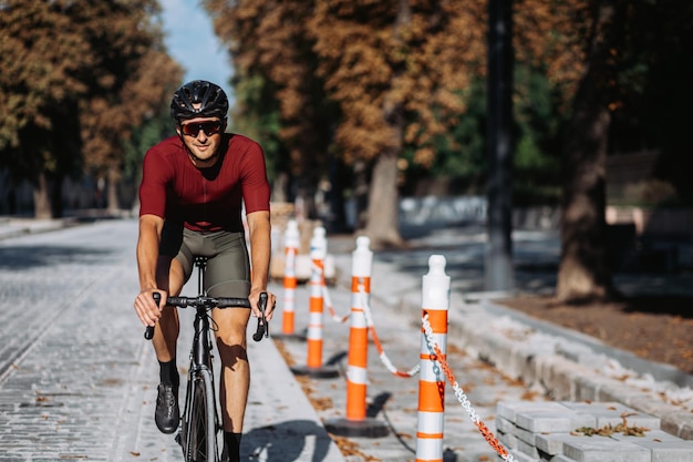 Homme caucasien positif en vêtements de sport, casque et lunettes passant le matin à faire du vélo dans la rue de la ville. Sportif bénéficiant d'entraînements réguliers en plein air.