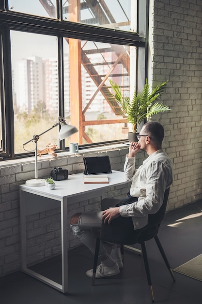 Un homme caucasien réfléchi regarde la fenêtre en travaillant au bureau avec la tablette et l'appareil photo