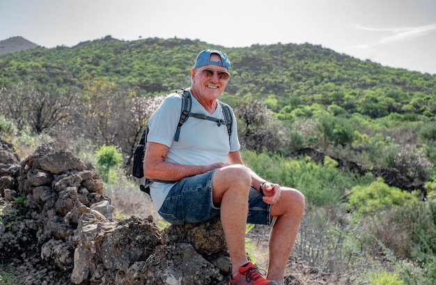 Un homme caucasien senior souriant avec une casquette et un sac à dos lors d'une excursion en montagne à l'extérieur s'assoit pour se reposer
