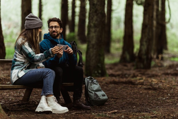 Un homme caucasien souriant du millénaire en veste montre un smartphone à sa femme s'asseoir sur un banc se détendre dans la forêt froide