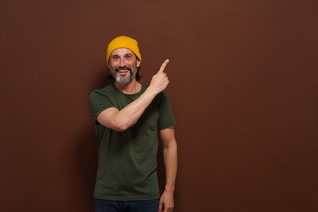 Photo un homme caucasien souriant et excité dans un chapeau jaune pointe sa main sur le côté avec un espace de copie sur un fond marron concept publicitaire de placement de produit