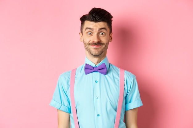 Homme caucasien souriant à la recherche d'excité, debout en noeud papillon élégant et chemise pour rendez-vous romantique, fond rose.