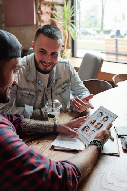 Homme célibataire assis à table dans un café et demandant conseil à un ami sur l'application de rencontres lors de la visualisation des profils de femmes sur tablette