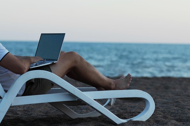 Un homme sur une chaise lounge sur la plage travaillant sur un ordinateur portable le concept de travailler en ligne n'importe où dans le monde