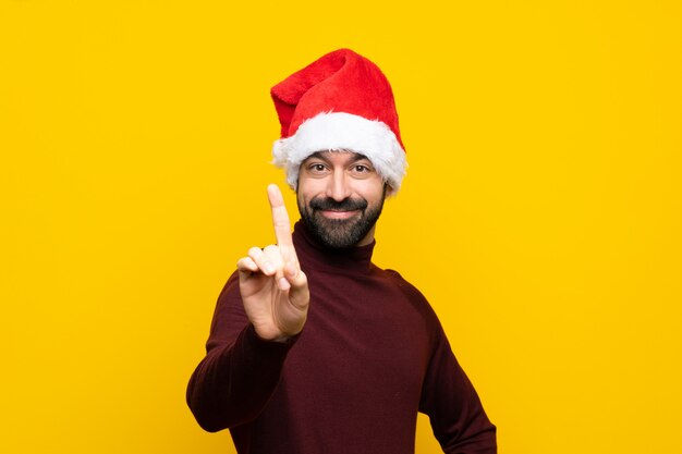 Homme avec chapeau de Noël sur mur jaune isolé montrant et en levant un doigt