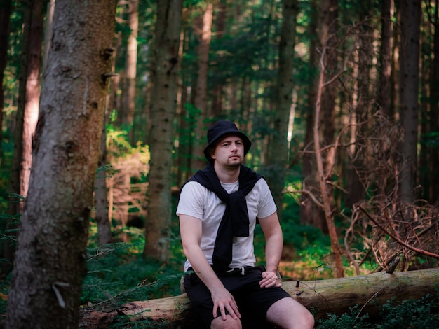 Un homme avec un chapeau noir et un t-shirt blanc est assis sur une bûche dans la forêt