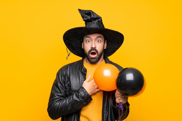 Homme avec chapeau de sorcière tenant des ballons à air noir et orange pour la fête d'halloween surpris et choqué tout en regardant à droite